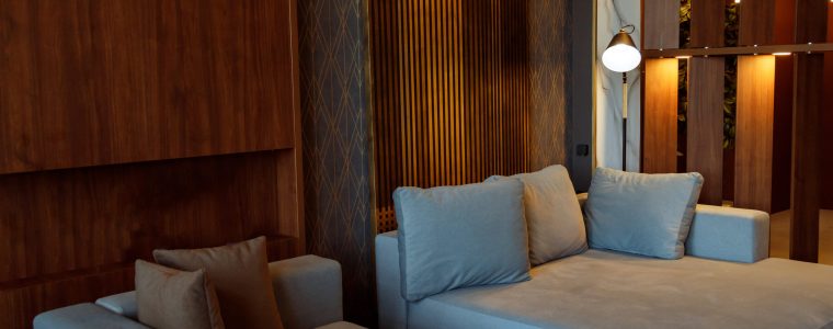 Modern interior design of living room with huge sofa, wooden planks on wall, golden ornate wallpaper, switched on floor lamp, night