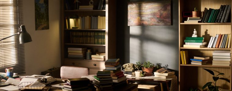 Sunlight streaming into home office filled with bookshelves, desk covered with stacked books, papers, potted plants and lamp, cozy workspace suggesting academic or creative activity